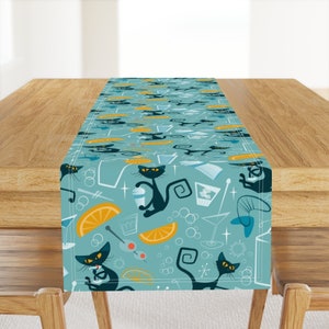 May include: A teal blue table runner with a repeating pattern of black cats, orange slices, and martini glasses. The pattern is in a retro style.
