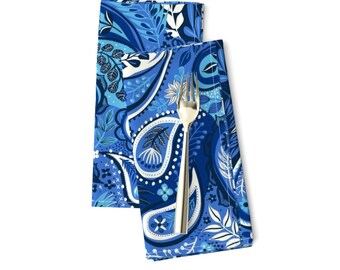 May include: Two blue paisley patterned cloth napkins with a silver fork. The napkins feature a variety of blue shades and white floral and leaf designs. The fork is placed on top of the napkins.