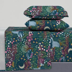 May include: A bedding set featuring a dark teal pattern with a whimsical forest design. The pattern includes trees, flowers, crescent moons, and stylized animals. Two pillowcases are stacked on top of the folded sheet.