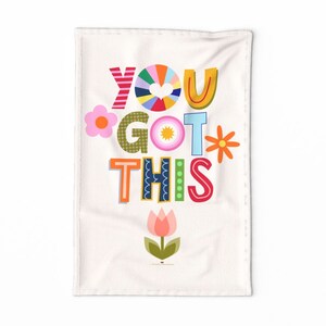 May include: A white kitchen towel with the text "YOU GOT THIS" in colorful, stylized letters. The letters are decorated with flowers, hearts, and dots. A pink tulip with green leaves is at the bottom of the towel.