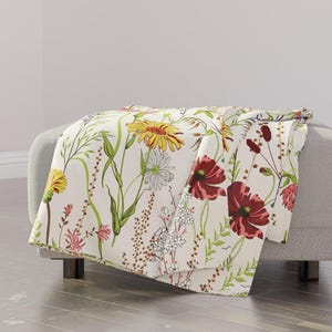 May include: A cream-colored throw blanket featuring a botanical print of various wildflowers, including large yellow sunflowers, deep red poppies, white daisies, and green grasses, arranged in a scattered, naturalistic pattern across the fabric.