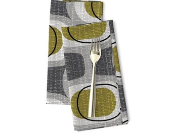 May include: Two folded cloth napkins with a mid-century modern design. The napkins feature a pattern of olive green and gray shapes outlined in black. A silver fork rests on top of the napkins.