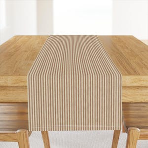 May include: A brown and cream striped table runner is centered on a wooden table. The runner has vertical stripes and is made of a woven fabric. The table and chairs are made of light-colored wood.