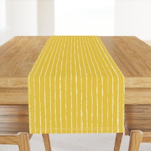 May include: A yellow table runner with a vertical white stripe pattern. The runner is placed on a wooden table, with the edges of the table and chairs visible. The design is simple and modern, suitable for home decor.