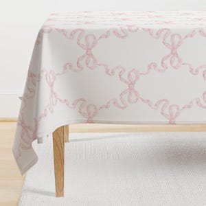 May include: A white tablecloth with a pink bow pattern. The tablecloth is draped over a table with wooden legs.