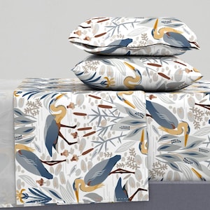 May include: A heron-themed bedding set. The set includes two pillows and a sheet, all patterned with blue, tan, and grey herons, leaves, and reeds on a white backdrop. The pillows are stacked on top of the sheet, creating a cohesive design.