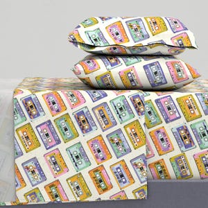 Puede incluir: Juego de cama con un patrón repetido de cintas de cassette coloridas sobre un fondo crema. El juego incluye una sábana encimera, una sábana bajera y dos fundas de almohada. Las cintas de cassette son en tonos amarillos, rosas, morados y verdes.