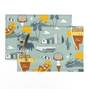 May include: Two placemats with a retro travel theme. The design features a light teal background with illustrations of palm trees, mountains, a vintage car, and a motel sign. The placemats have a fork and knife on top.