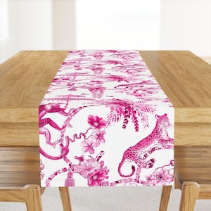 May include: Pink table runner with a white background featuring a repeating pattern of pink leopards, monkeys, and flowers.