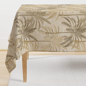 Op de afbeelding: Een beige tafelkleed met een bruin palmenmotief. Het tafelkleed is over een houten tafel gelegd.