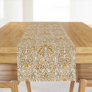 May include: A gold and white patterned table runner. The runner features an intricate floral design with gold and white accents. The runner is displayed on a wooden table, adding a touch of elegance to the dining area.