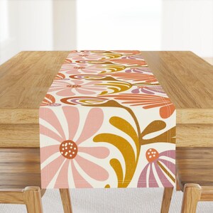 May include: A floral table runner with a retro design. The runner features large pink and orange flowers with brown and yellow accents on a cream background. The runner is displayed on a wooden table.