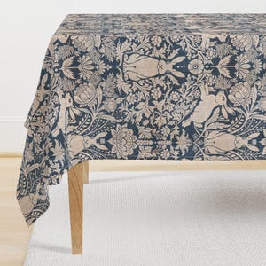 May include: A rectangular tablecloth with a navy blue background and a beige floral and rabbit pattern. The tablecloth is draped over a wooden table, with the corner of the tablecloth hanging down. The table is set on a white rug.
