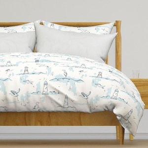 May include: White duvet cover with a light blue and brown pattern of lighthouses, birds, and clouds. The duvet cover is on a bed with a wooden headboard and a white pillow.