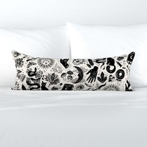 May include: A long, rectangular pillow with a cream-colored background and a repeating black pattern of mystical symbols. The design includes moons, suns, snakes, hands, crystals, and mushrooms. The pillow is placed on a white bed.