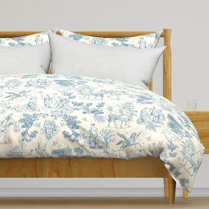 Op de afbeelding: Een witte dekbedovertrek met een blauw toile-patroon met verschillende afbeeldingen, waaronder bomen, vogels en mythische wezens. De dekbedovertrek ligt op een bed met een houten hoofdeinde en een wit kussen.
