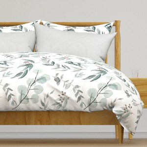 May include: A bed with a wooden frame and white pillows. The bedding features a botanical pattern of green and grey eucalyptus leaves and berries on a white background. The bed is in a well-lit room.
