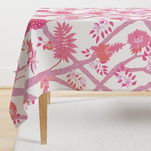 Puede incluir: Un mantel blanco con un patrón floral rosa. El patrón presenta flores rosas y naranjas, hojas y ramas. El mantel está drapeado sobre una mesa con patas de madera.