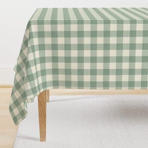 May include: A sage green and cream gingham tablecloth on a wooden table. The checkered pattern creates a classic, country-style aesthetic. The tablecloth drapes over the edges of the table, showcasing the fabric's texture and design. The table is approximately 100 cm high.