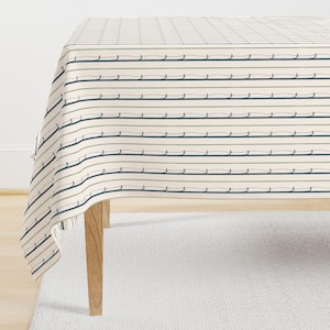 May include: Cream-colored tablecloth with a repeating pattern of horizontal lines in navy blue, olive green, and light gray. The lines are interspersed with a wavy design. The tablecloth is draped over a wooden table.