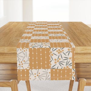 Könnte beinhalten: Rechteckiger Tischläufer mit Patchwork-Design. Der Läufer zeigt Quadrate in Creme, Senfgelb und Weiß mit floralen und geometrischen Mustern. Der Läufer liegt auf einem Holztisch.