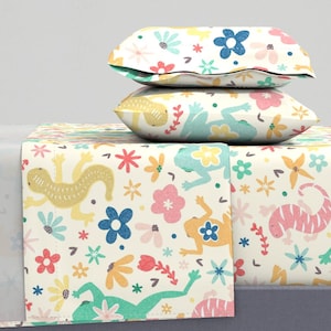 May include: A cream-colored bedding set featuring a whimsical floral and animal print. The set includes a flat sheet, fitted sheet, and two pillowcases. The print showcases colorful flowers and playful lizards and frogs in shades of yellow, pink, blue, and green.