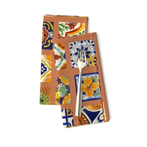 May include: Two orange napkins with a colorful tile pattern. The tiles are in shades of blue, green, yellow, and red. The napkins are folded and a silver fork is placed on top.