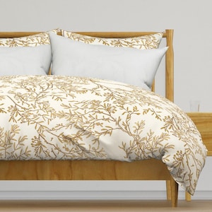 May include: A white duvet cover with a gold floral pattern featuring delicate branches and leaves. The duvet cover is on a wooden bed frame with a white pillow.