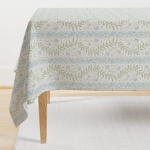May include: A white tablecloth with a delicate floral pattern in shades of blue and green. The pattern features small flowers and leaves arranged in a repeating design. The tablecloth is shown draped over a wooden table.