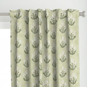 Peut inclure: Rideaux à imprimé floral vert clair. Les rideaux présentent un motif répété de grappes de fleurs blanches avec des feuilles vertes. Le tissu est une matière douce et texturée, et les rideaux sont suspendus à une tringle à rideaux argentée.