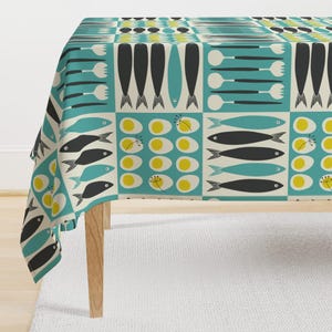 May include: A rectangular tablecloth with a repeating pattern of black fish, yellow and white eggs, and teal utensils on a cream and teal background. The tablecloth is draped over a wooden table.