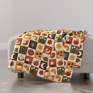 Op de afbeelding: Een sprei met een herhalend patroon van herfstthema-vierkanten. Elke vierkant bevat illustraties van appels, bladeren, paddenstoelen en andere herfstelementen in tinten oranje, bruin, groen en crème. De sprei is gedrapeerd over een grijze poef.