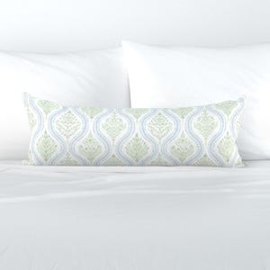 May include: A long, rectangular decorative pillow with a repeating pattern of stylized floral designs in light green and blue on a white background. The pillow is placed on a white bed with white pillows.