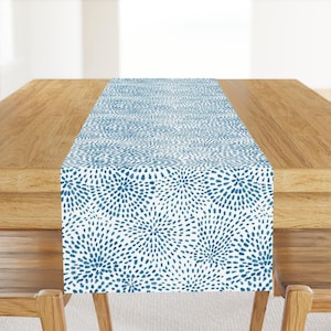 Puede incluir: Un camino de mesa azul y blanco con un patrón repetitivo de explosiones florales estilizadas. El camino de mesa se muestra sobre una mesa de madera, mostrando su diseño y textura. Esta pieza decorativa añade un toque de elegancia a cualquier comedor.