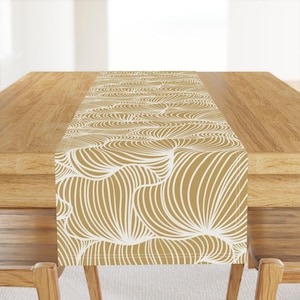 May include: A gold and white table runner with a wavy, abstract pattern. The runner is made of fabric and has a subtle sheen.