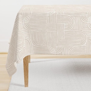 May include: A beige tablecloth with a repeating pattern of white, curved lines. The lines create an abstract design of arches and rectangles. The tablecloth is draped over a wooden table, with a white rug visible beneath.
