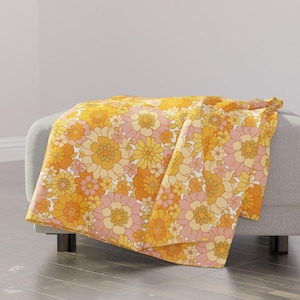 May include: A yellow and pink floral throw blanket with a retro 70s style pattern. The blanket is folded and draped over a light gray couch.