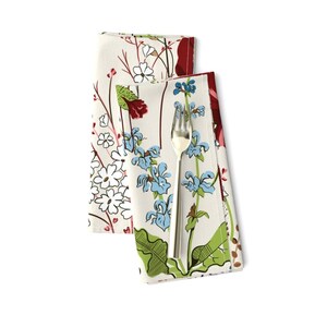 May include: A folded white fabric napkin featuring a botanical print of blue, red, and white wildflowers with green stems and leaves. A silver-colored metal fork with three tines is placed vertically on top of the napkin, centered on the floral pattern.