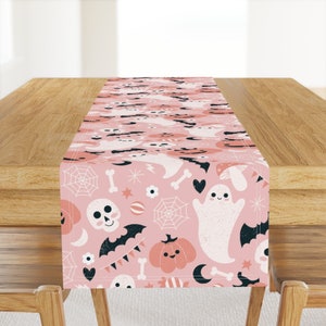 May include: A pink table runner with a Halloween theme featuring ghosts, pumpkins, bats, skulls, and candy corn.