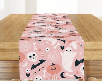 May include: A pink table runner with a Halloween theme featuring ghosts, pumpkins, bats, skulls, and candy corn.