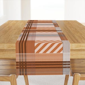 Op de afbeelding: Een rechthoekige tafelloper met een geometrisch patroon in bruin-, oranje- en wittinten. Het ontwerp heeft strepen, lijnen en een ruitvormige opstelling. De loper ligt op een houten tafel.