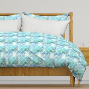 May include: A light blue and white duvet cover with a mermaid scale pattern. The duvet cover is on a wooden bed frame with white pillows.