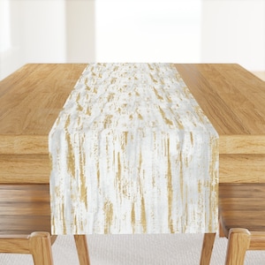 May include: White table runner with a gold abstract pattern. The runner is on a wooden table.