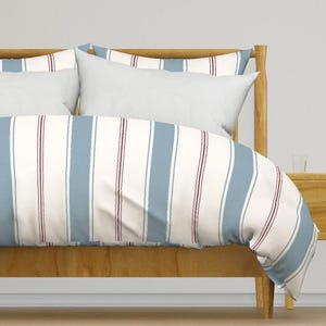Pode incluir: Um conjunto de roupa de cama com capa de edredão e almofadas com um padrão de riscas verticais em azul poeirento, creme e linhas finas vermelho-escuras. A roupa de cama está sobre uma estrutura de cama de madeira castanho-clara.
