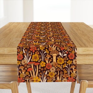 May include: A brown table runner with a vintage floral and mushroom pattern. The pattern features orange, yellow, and white flowers, brown mushrooms, and green leaves on a dark brown background.