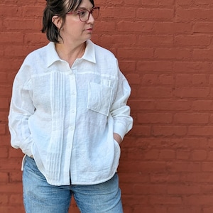 May include: A person wearing a white linen shirt with pleat detailing, blue jeans, and black sneakers. The shirt has a button-down front and a chest pocket. The jeans are cuffed at the ankles. The background is a red brick wall.
