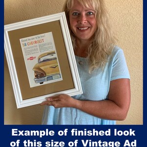 May include: A framed vintage advertisement for a 1958 Chevrolet car. The ad features a yellow car with red and white text. The frame is white, and the matting is tan. The image is held by a person.
