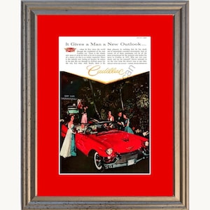 May include: A framed vintage advertisement for a Cadillac car featuring a red convertible, people in formal attire, and the text It Gives a Man a New Outlook. The print is set against a bright red matting inside a textured silver-toned frame.