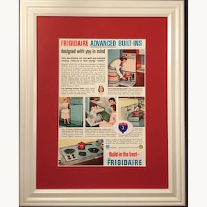 Può includere: Una pubblicità vintage Frigidaire Advanced Built-ins incorniciata in una cornice di legno bianco con un passe-partout rosso. La stampa mostra elettrodomestici da cucina di metà secolo, tra cui un frigorifero, una lavastoviglie e un fornello.