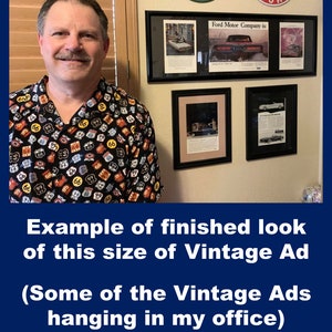 May include: A man wearing a Route 66 themed shirt stands in front of framed vintage car advertisements. The ads are in black frames and are mounted on a beige wall. Above the frames are vintage gasoline and motor oil signs.